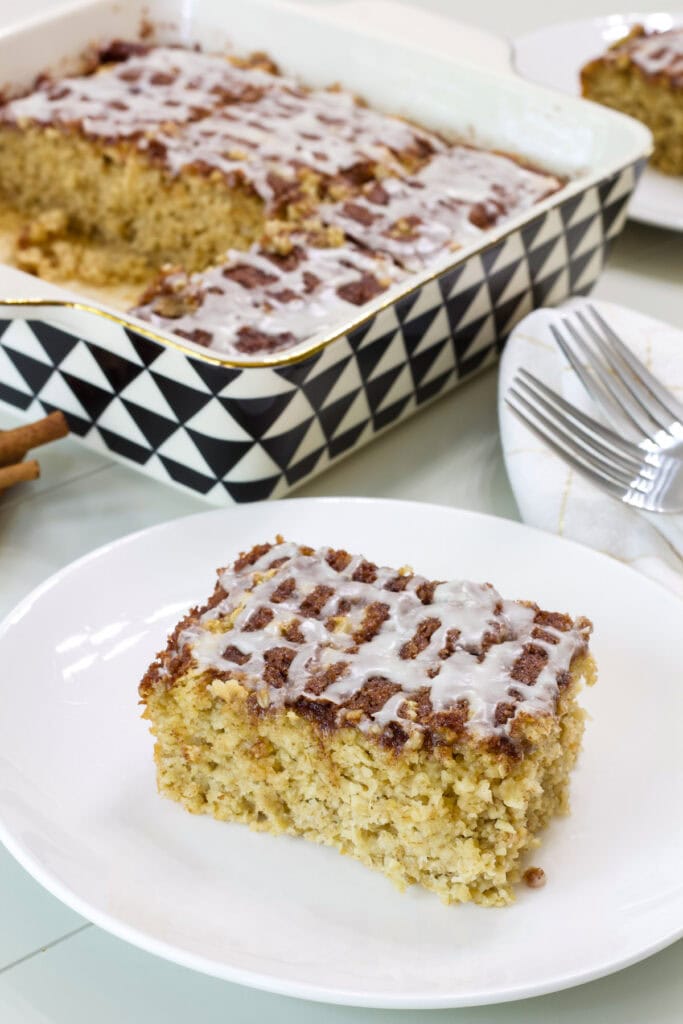 A piece of cinnamon roll baked oatmeal on a white plate and the pan with the remaining in the background.