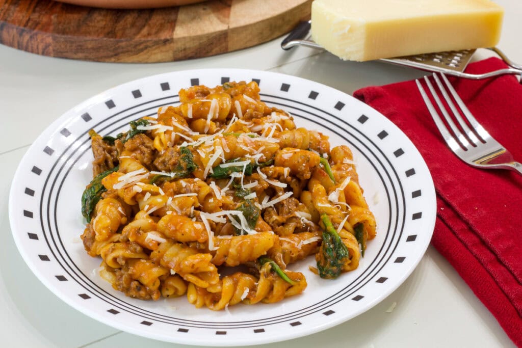 One serving of One Pot Ground Beef Pasta on a plate with a napkin, fork, and wedge of cheese next to it.