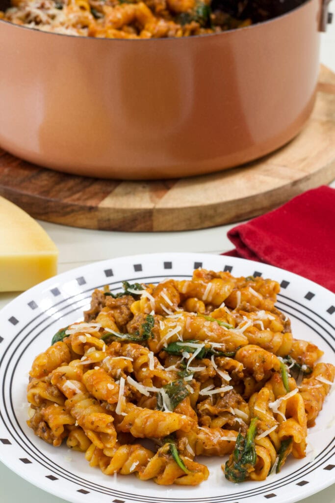 One serving of One Pot Ground Beef Pasta on a plate in the foreground and a pan with pasta in it in the background.