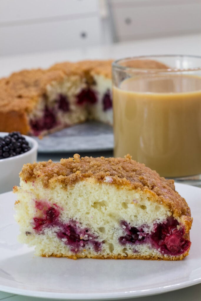 A slice of black berry coffee cake with streusel topping in the foreground with a cup of coffee and the remaining cake in the background.