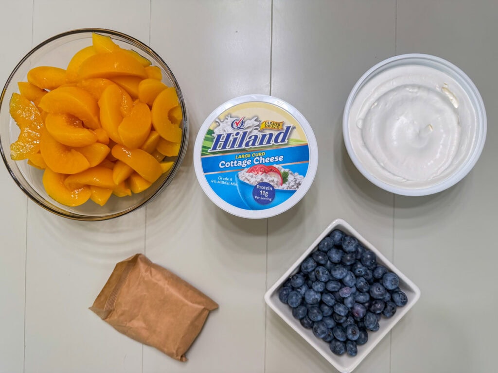 A bowl of sliced peaches, containers of cottage cheese and whipped topping, a package of vanilla pudding and a basket of fresh blueberries.
