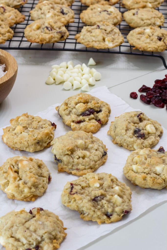 Several cowgirl cookies on a piece of parchment paper with white chocolate chips and dried cranberries in the background.