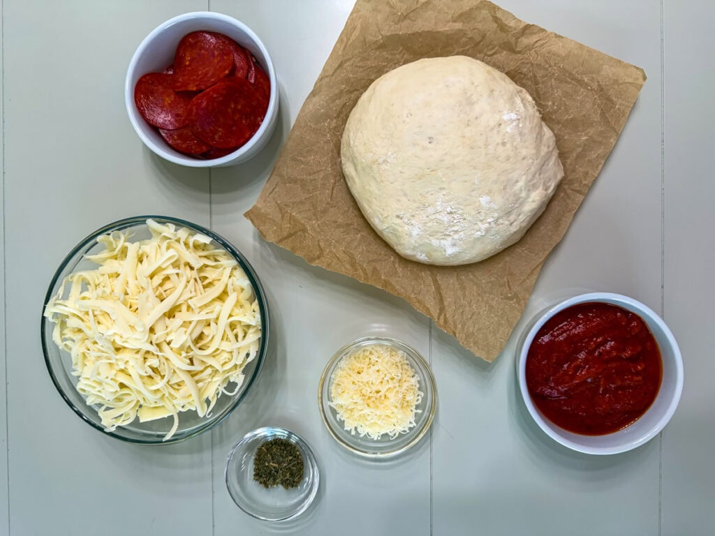 A ball of pizza dough, pepperoni, mozzarella cheese, parmesan cheese, pizza sauce and Italian seasoning needed to make a homemade pepperoni pizza.