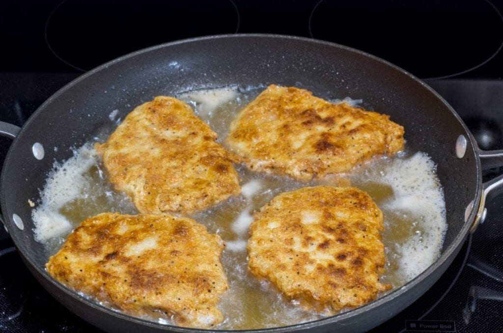 Four breaded pork chops frying in oil in a large skillet.