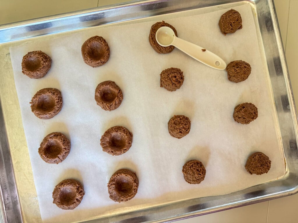 The cookie dough balls on a parchment paper lined baking sheet, some have been indented and some have not.