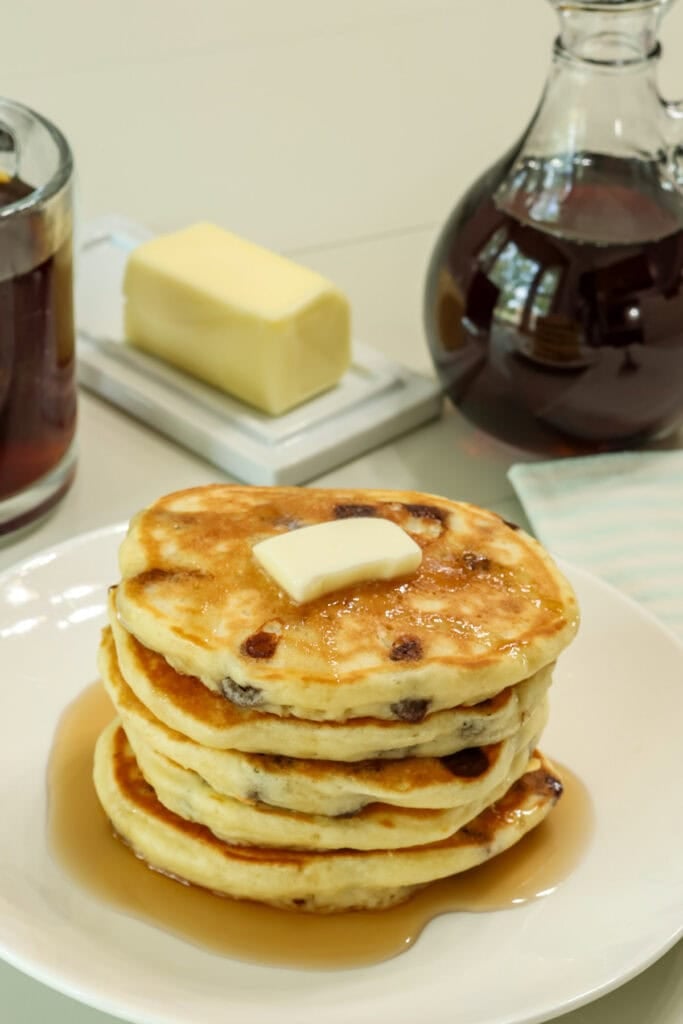 A stack of fluffy chocolate chip pancakes topped with butter and syrup on a white plate.