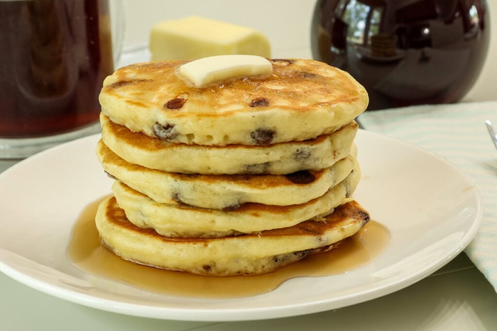 Closeup side view of a stack of five fluffy chocolate chip pancakes topped with a pat of butter and pancake syrup.