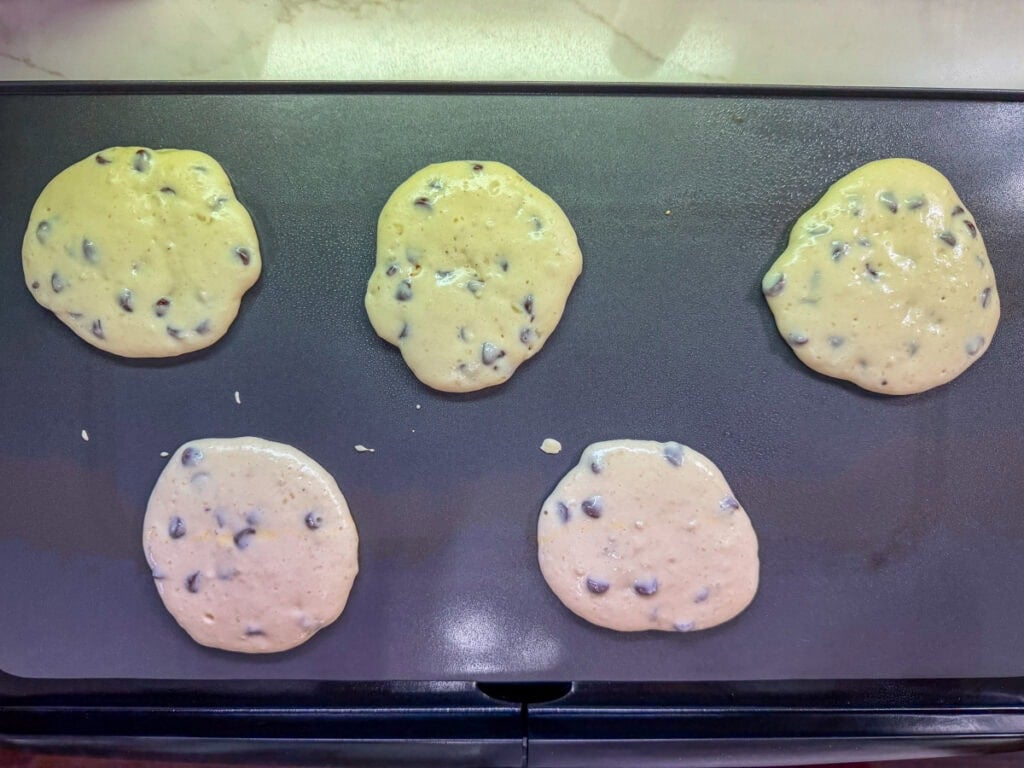 Five chocolate chip pancakes on a griddle before being flipped over.
