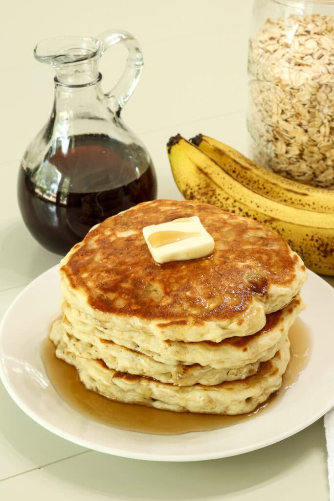 A stack of four banana oatmeal pancakes and oats, bananas and pancake syrup in the background.