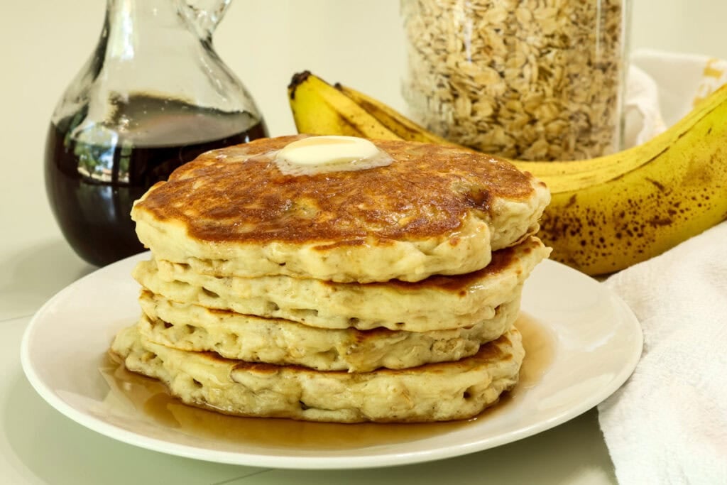 Side view of a stack of four banana oatmeal pancakes topped with a pat of butter and syrup.