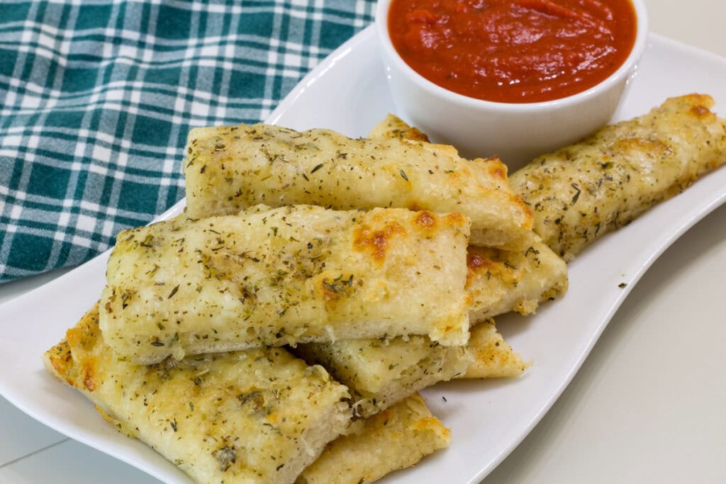 Side view of several pizza dough breadsticks on a white plate with a small bowl of marinara sauce.