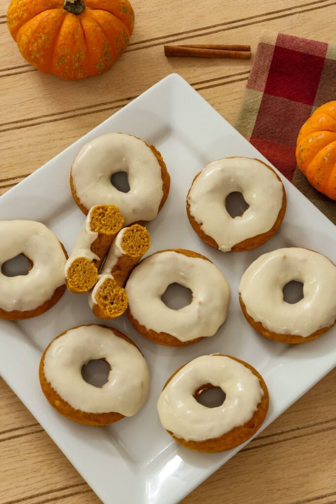 Eight baked pumpkin donuts on a white plate with one cut in half so the tender inside is visible.