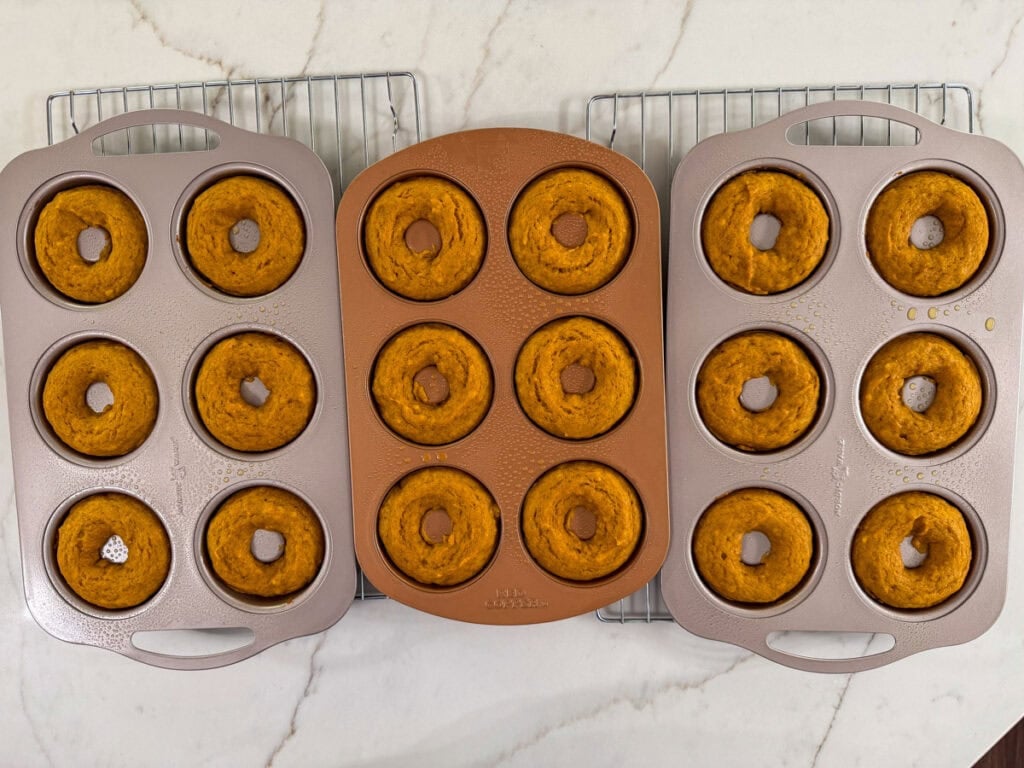 Three donut pans with baked donuts in the on wire cooling racks.