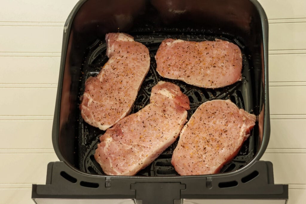 Four seasoned uncooked pork chops in an air fryer basket.